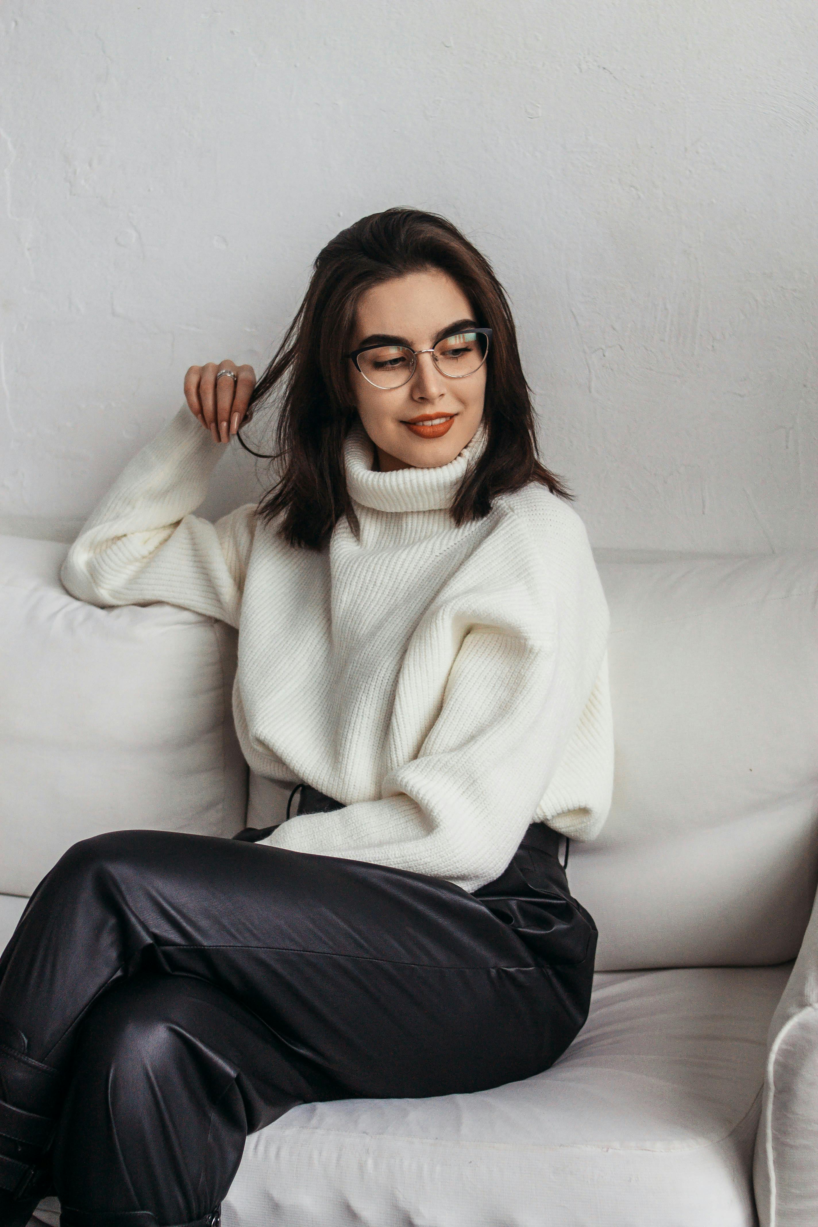 Chic woman in glasses wearing a white turtleneck sweater while sitting on a sofa.