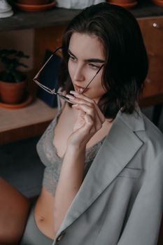 Alluring woman with dark hair and eyeglasses wearing stylish lingerie and gray blazer while sitting in room near potted plants
