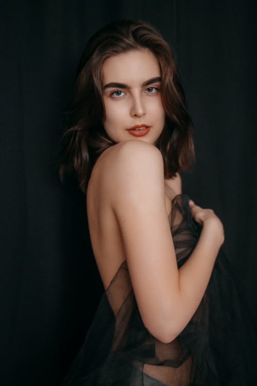 Sensual young topless female with dark hair in panties covering with textile and looking at camera against black background in studio