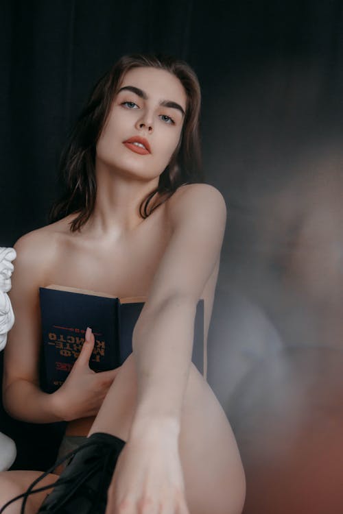 Free Sensual slim naked female in boots covering breast with book and looking at camera while sitting on black background in studio Stock Photo
