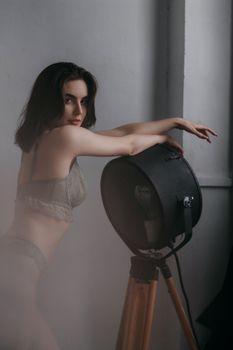 Alluring slender young female with dark hair in lingerie looking at camera while leaning on lightning equipment in light studio
