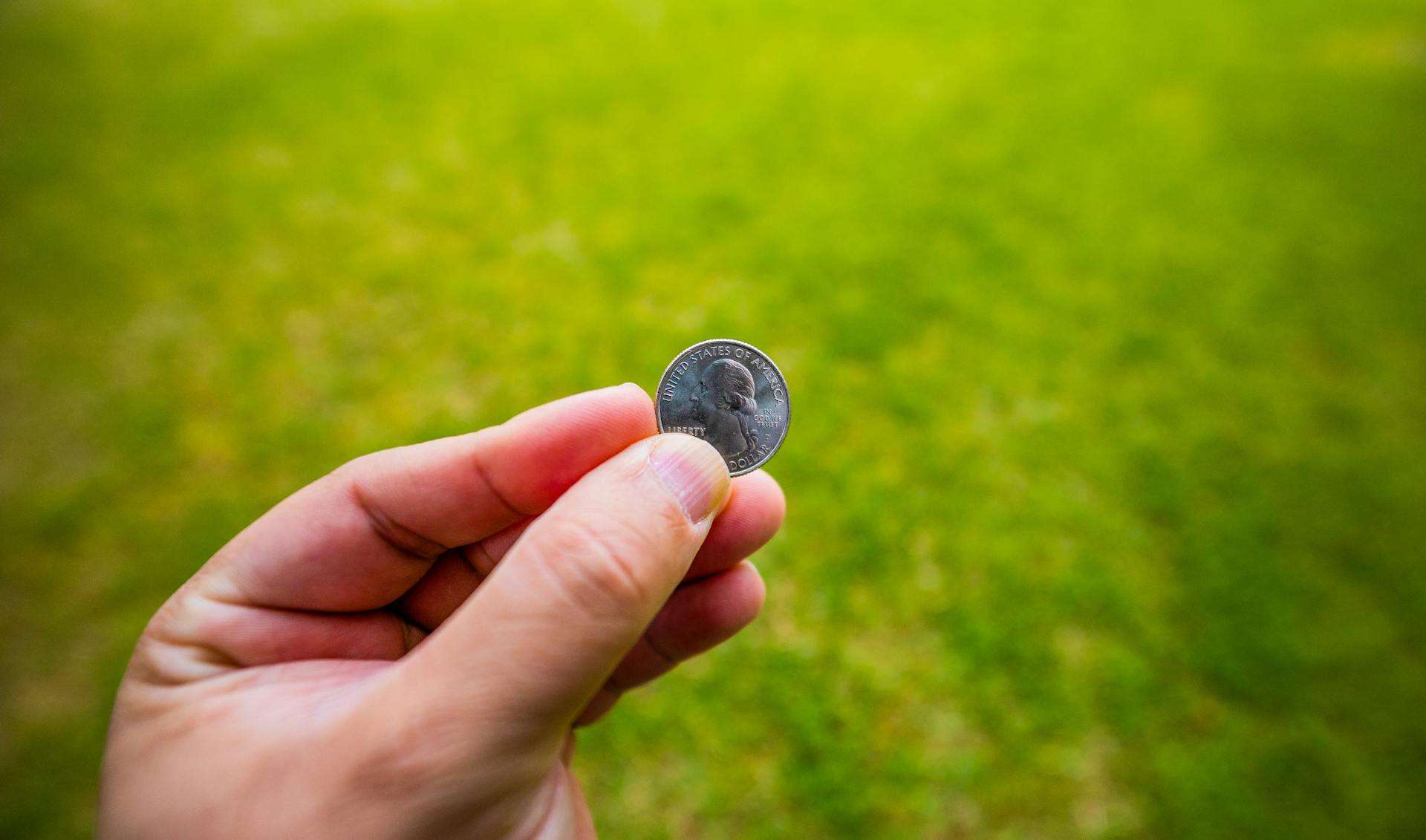 Vibrant close-up shot of a hand holding a small silver coin against a blurred green grass background.