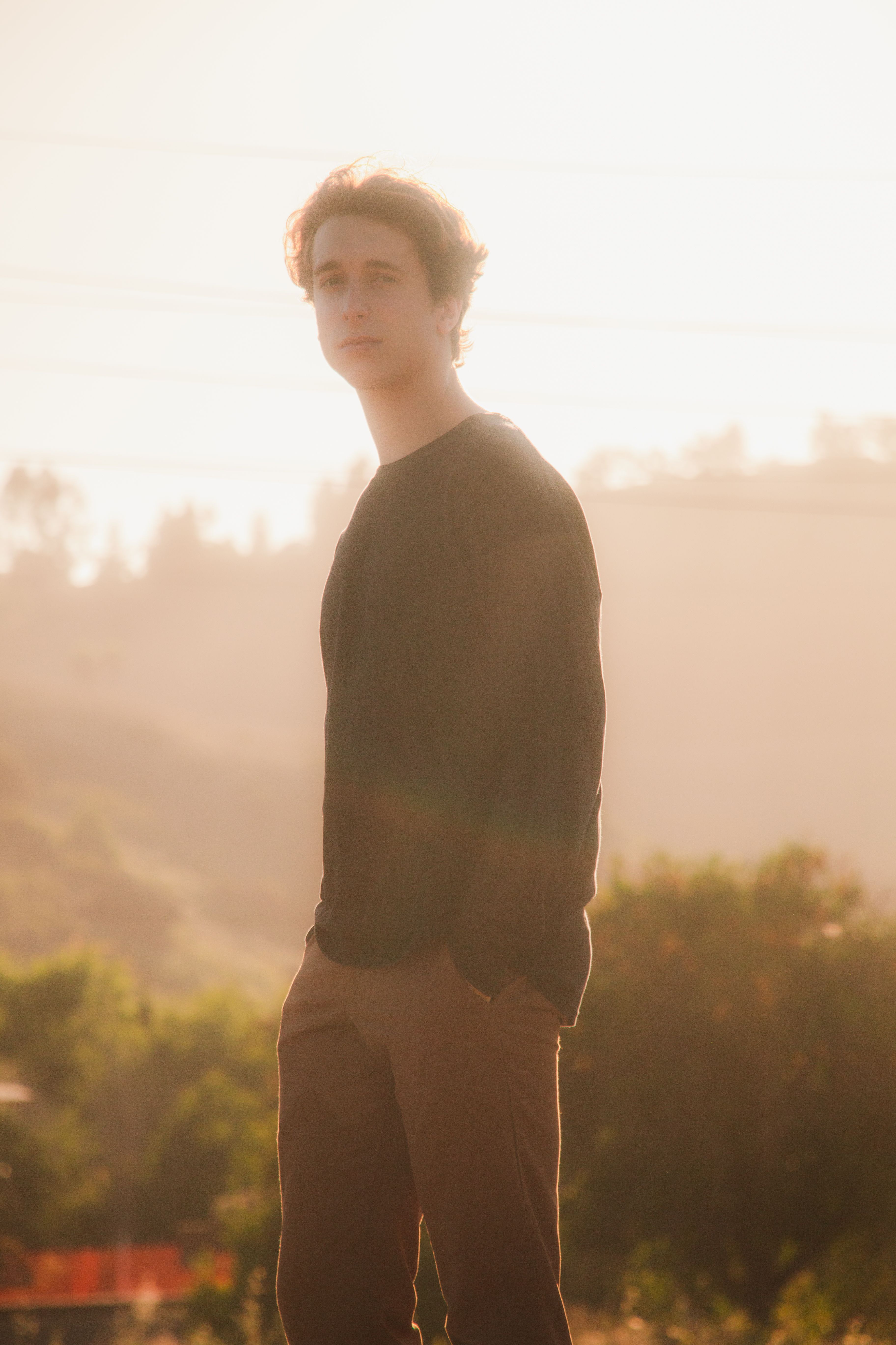 A young man stands outdoors in casual clothing, captured in a serene, sunlit setting.