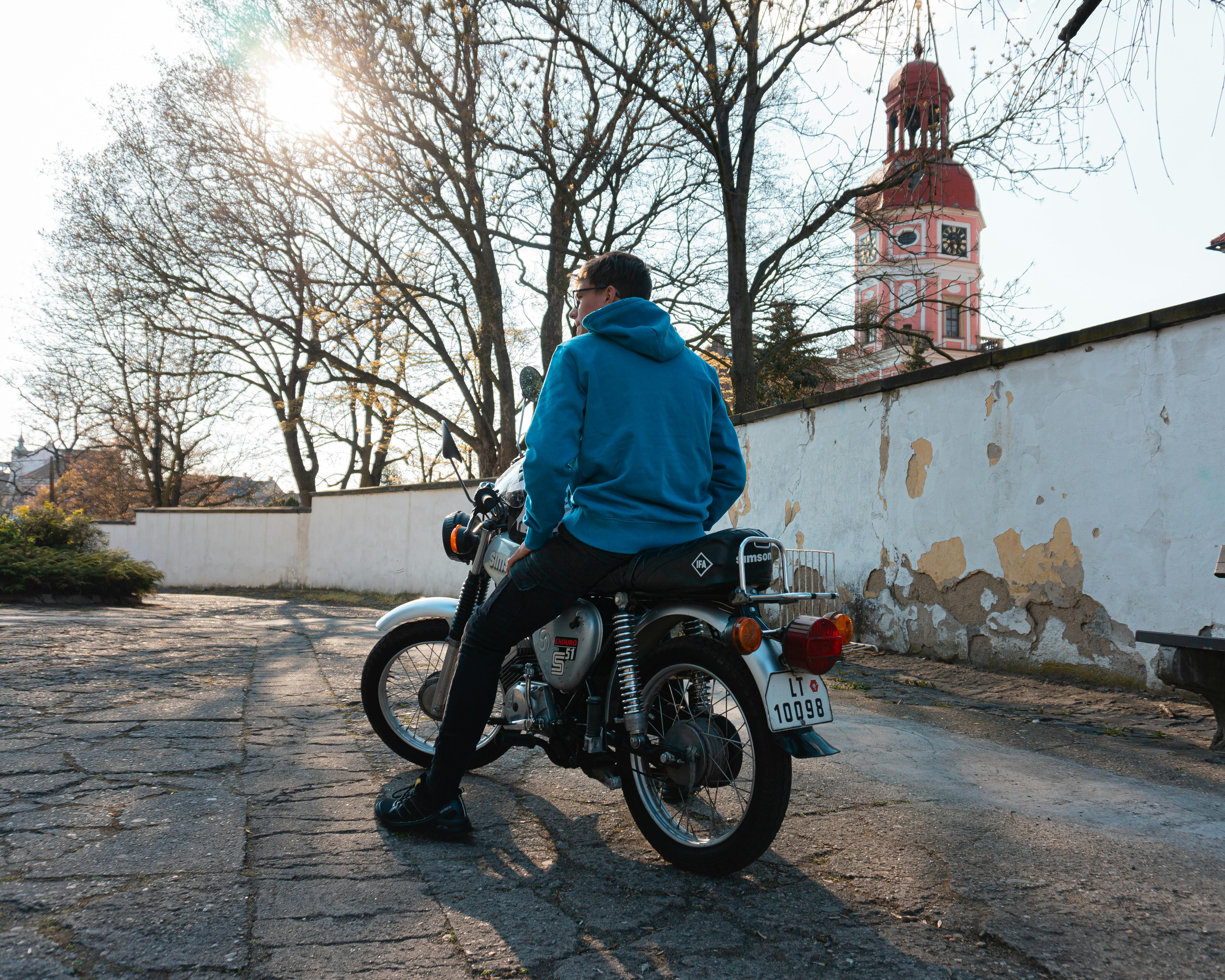 A man in a blue hoodie sits on a motorcycle in an urban environment with a clock tower.