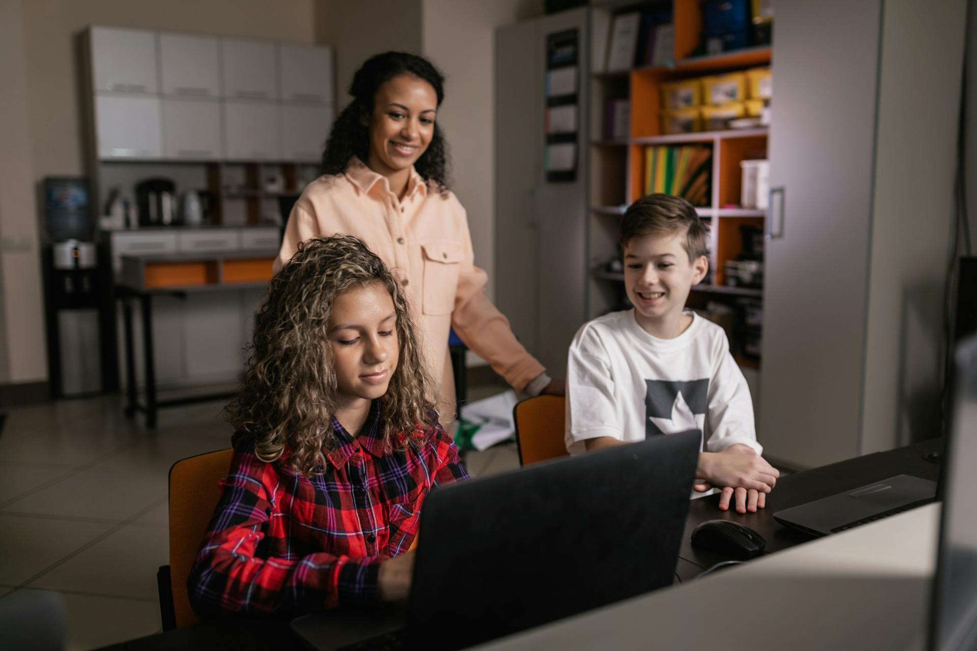 A teacher supervising two smiling teenagers working on laptops in a classroom setting, focusing on technology and learning.