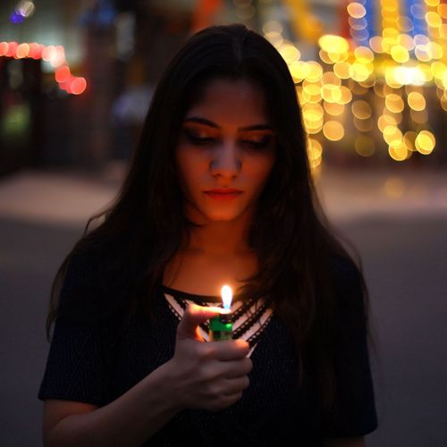 Free Young woman holding a lighter with colorful bokeh lights in the background, creating a moody urban portrait. Stock Photo