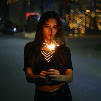 A young woman holding a bright sparkler outdoors at night with bokeh lights around.