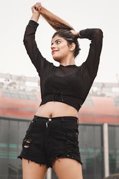 Confident young woman in black crop top with ripped shorts holding her hair outdoors.