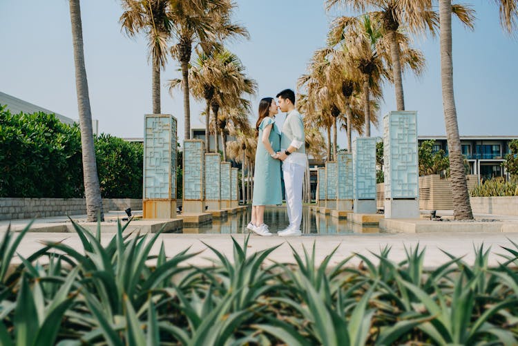 Loving Asian Couple Holding Hands Near Fountain In Modern Resort