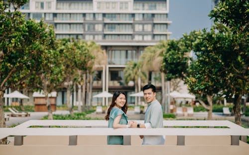 Side view cheerful young Asian couple holding hands and looking at camera while standing in sunny city park