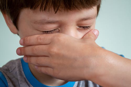 Crop little boy with closed eyes and brown hair crying and wiping tears from cheeks against white background
