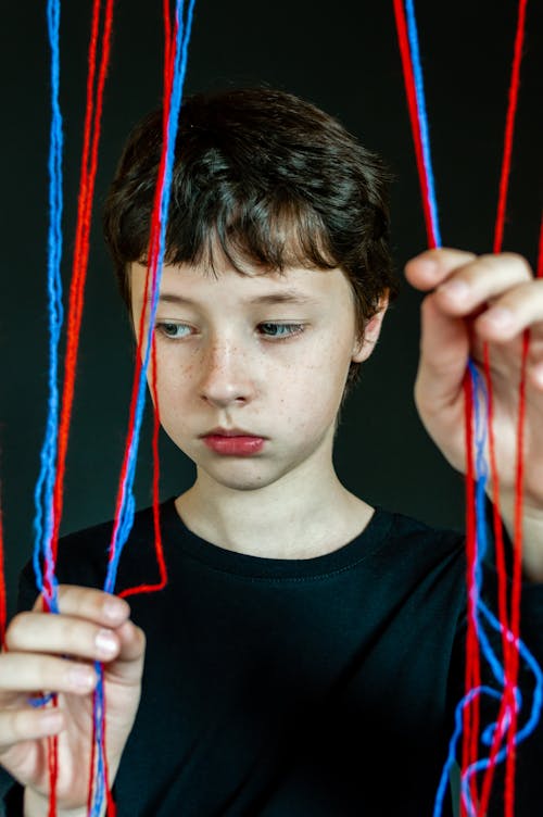 Unemotional child with brown hair in black t shirt standing against black background and holding red and blue threads while looking down