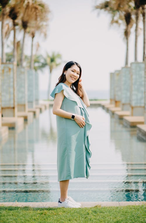 Side view of cheerful young ethnic pregnant female in trendy outfit adjusting hair and touching tummy while smiling and looking at camera near swimming pool in tropical resort