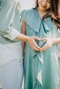 Crop happy pregnant Asian woman with anonymous husband doing heart with hands near tummy on sunny day