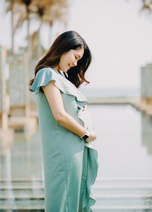 Side view of happy young ethnic pregnant lady with long dark hair in elegant dress smiling and caressing tummy while standing at poolside in tropical resort