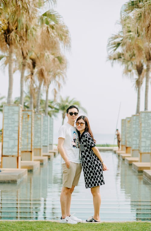 Full body of couple in sunglasses standing near open pool with palm trees in summer park while looking at camera
