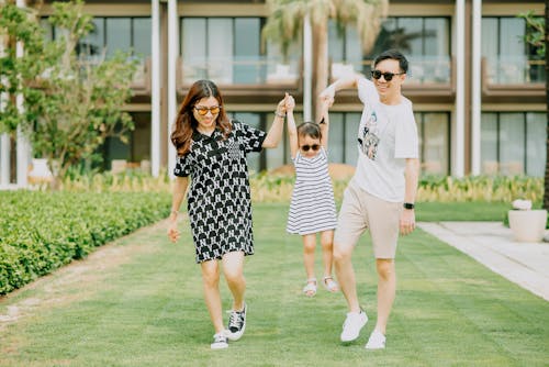 Free Cheerful young Asian mother and father wearing casual clothes and laughing while raising daughter on hands and standing on green lawn in front of resort territory Stock Photo