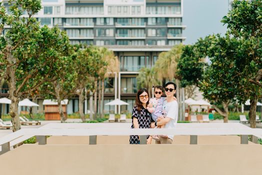 Positive young Asian couple with child wearing casual clothing and stylish sunglasses while standing among green tropical trees and looking at camera