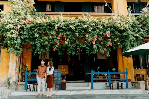 Full body of young romantic Asian couple wearing casual clothes and standing together near entrance of cafe decorated with blooming greenery