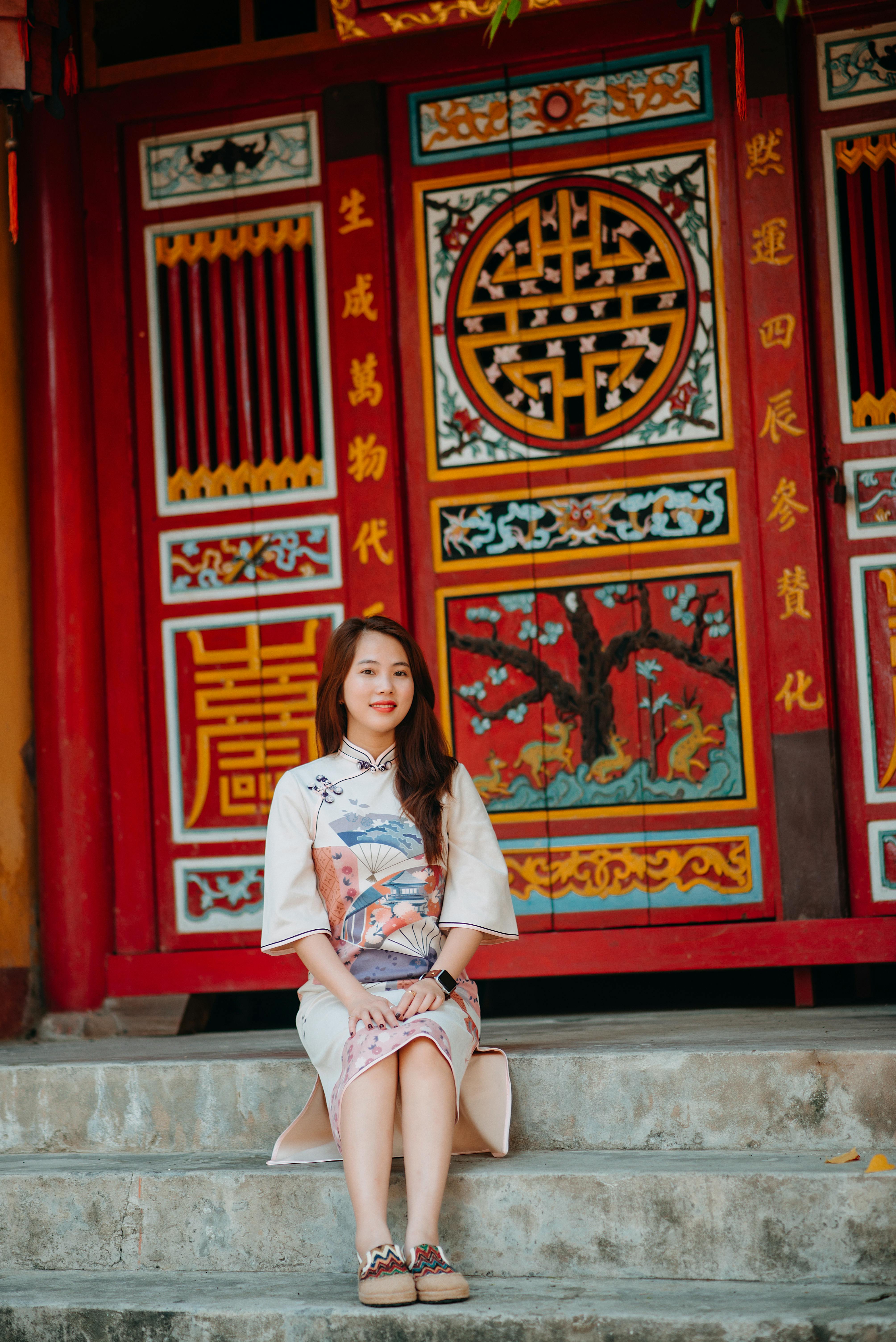 Full body of positive young ethnic lady in elegant dress smiling and looking at camera while sitting in stair of oriental temple