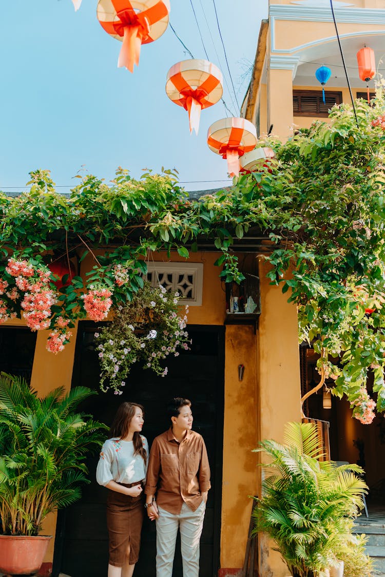 Fashionable Asian Couple Holding Hands And Looking Away Near Mansion And Exotic Plants