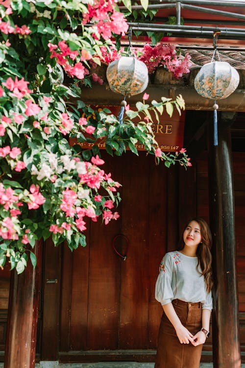 Free Romantic young Asian lady with long hair in casual clothes leaning on pillar of wooden house and smiling while admiring blooming tree on sunny day Stock Photo