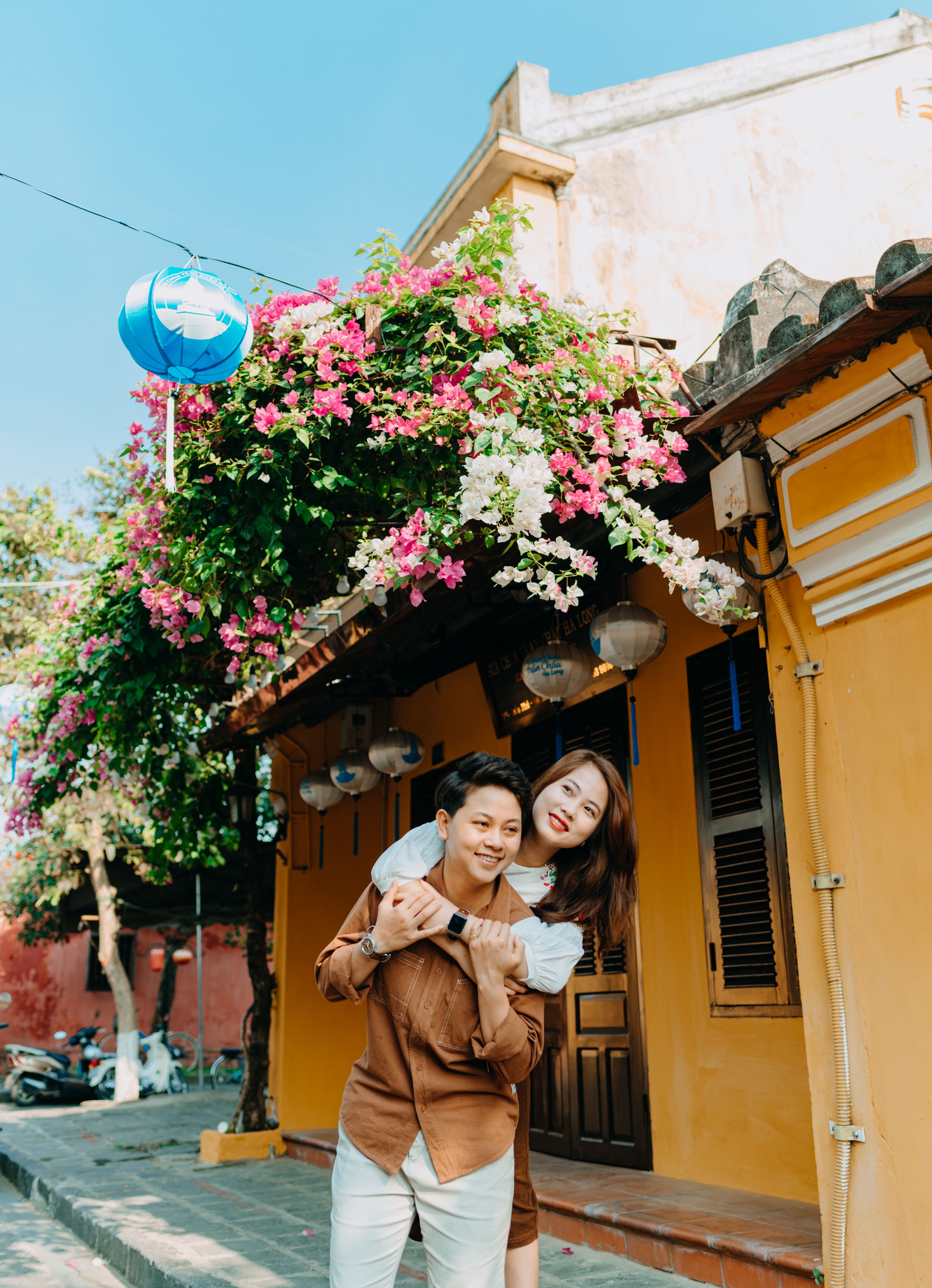 Happy couple embracing joyfully on a colorful street with flowers overhead.