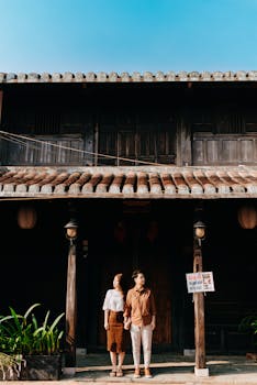 Full length of young ethnic couple in stylish clothes holding hands and looking in different directions while standing together near aged wooden house on sunny day