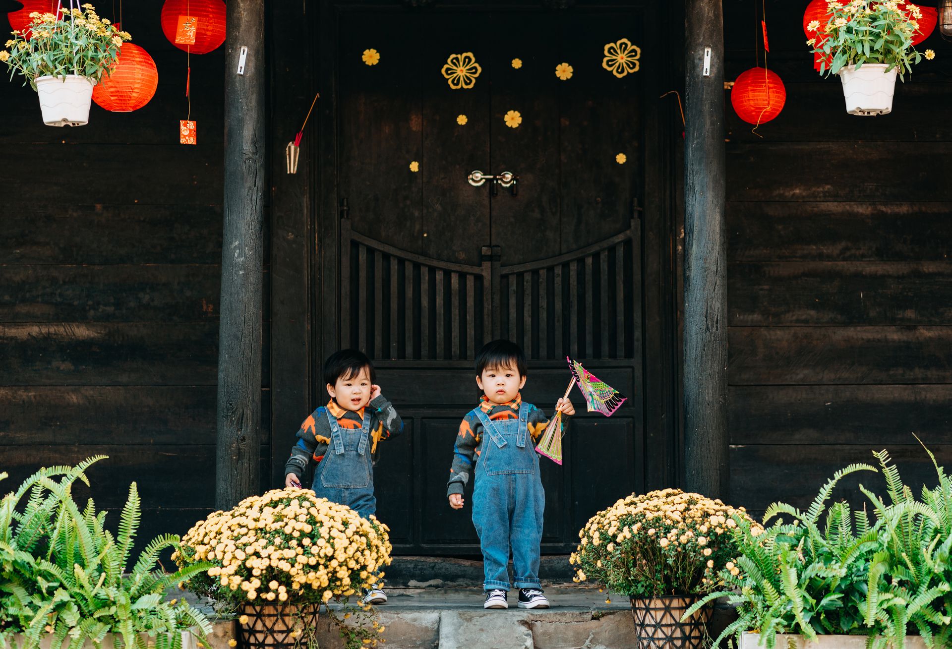 Full length of adorable ethnic twin toddlers in similar trendy denim outfits standing near wooden house amidst blooming plants before launching kite in park