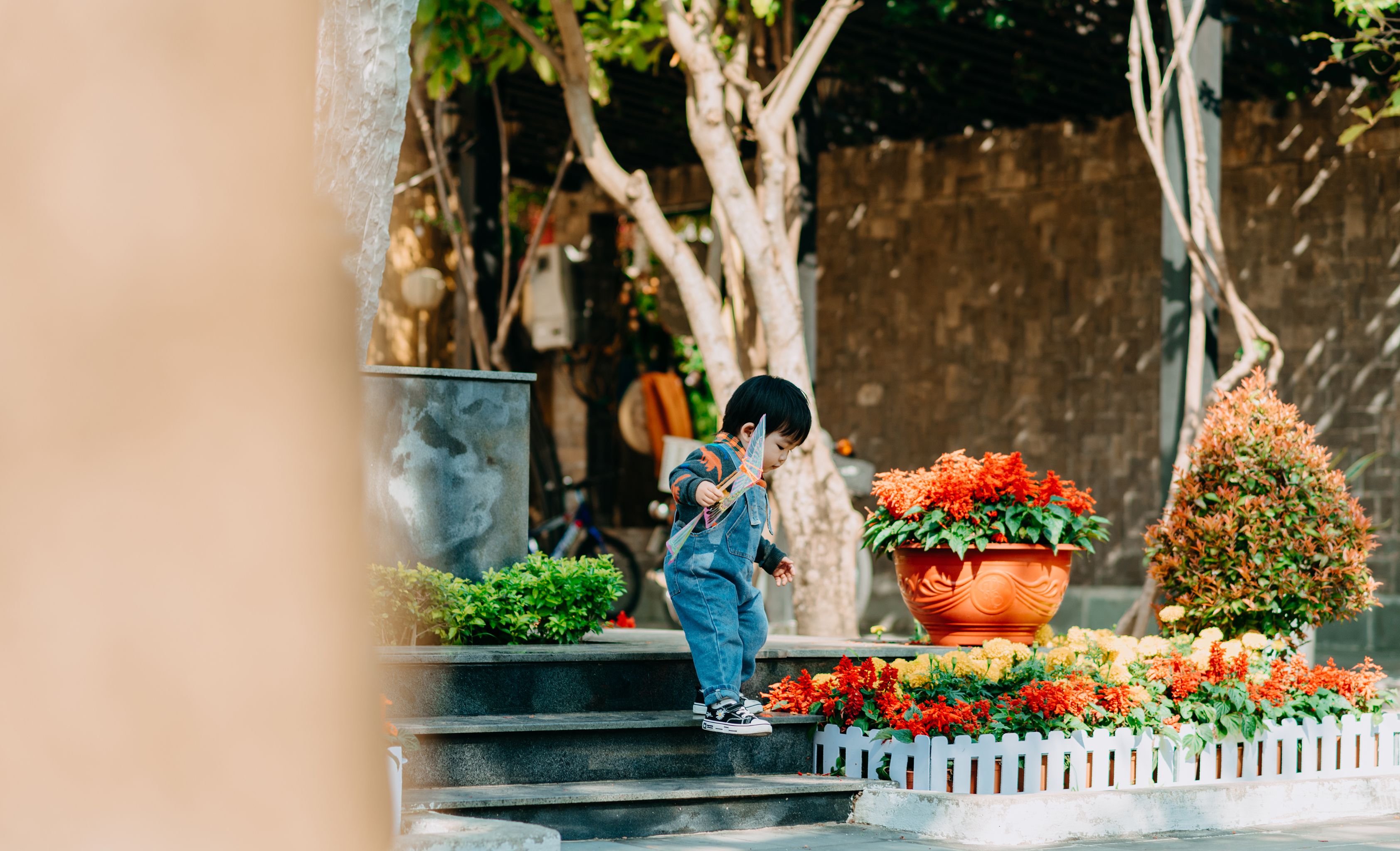 Side view of adorable curious little ethnic boy in trendy clothes walking down steps near potted blooming plants on sunny day in park