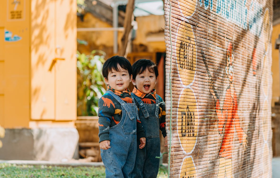 Adorable little Asian twin brothers in similar denim overalls smiling and looking at camera while standing near bamboo wall in backyard on sunny day Stock Photo