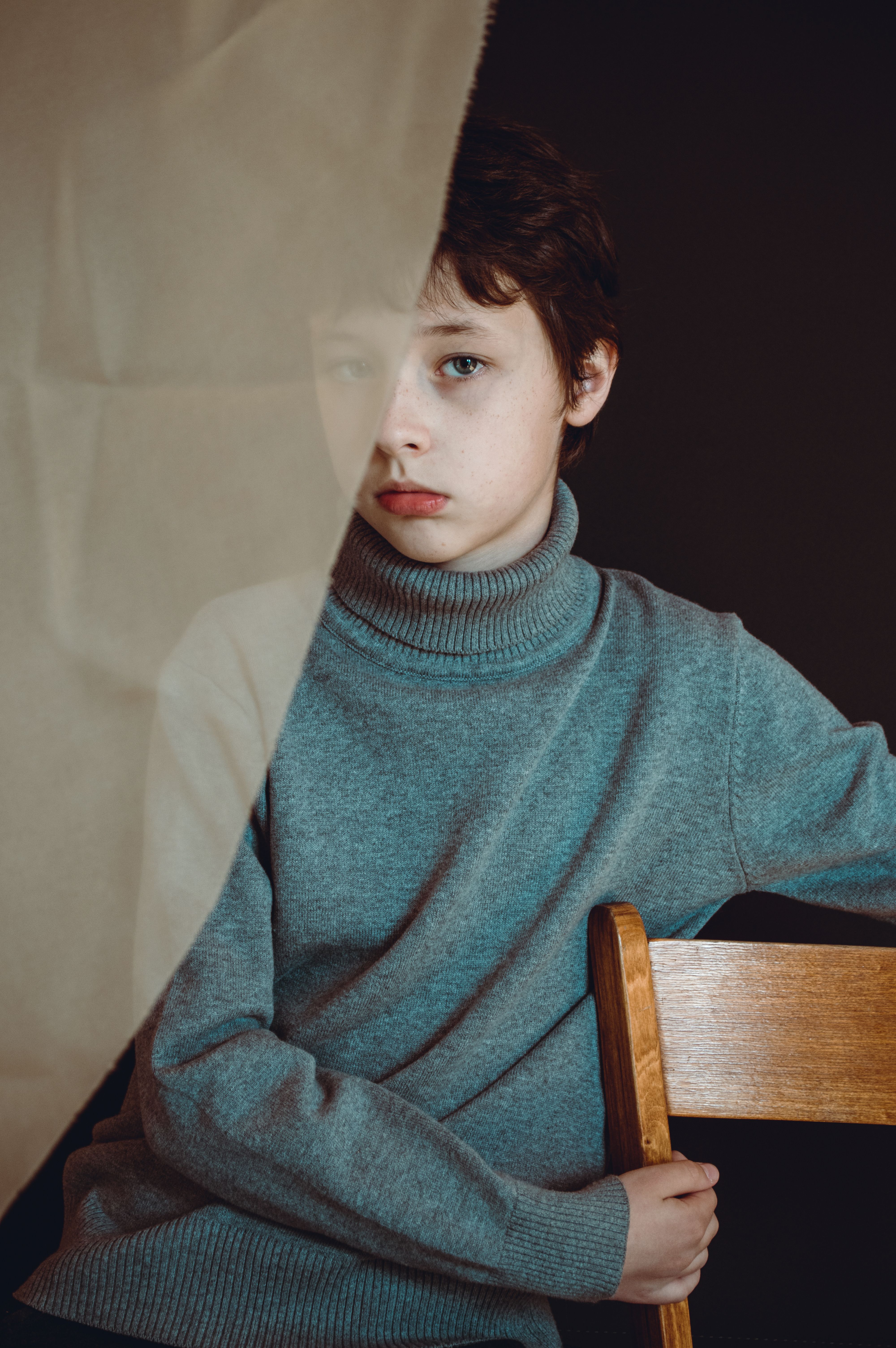 Unemotional male child with brown hair wearing warm gray turtleneck sweater covered with transparent curtain sitting on wooden stool against black background and looking at camera