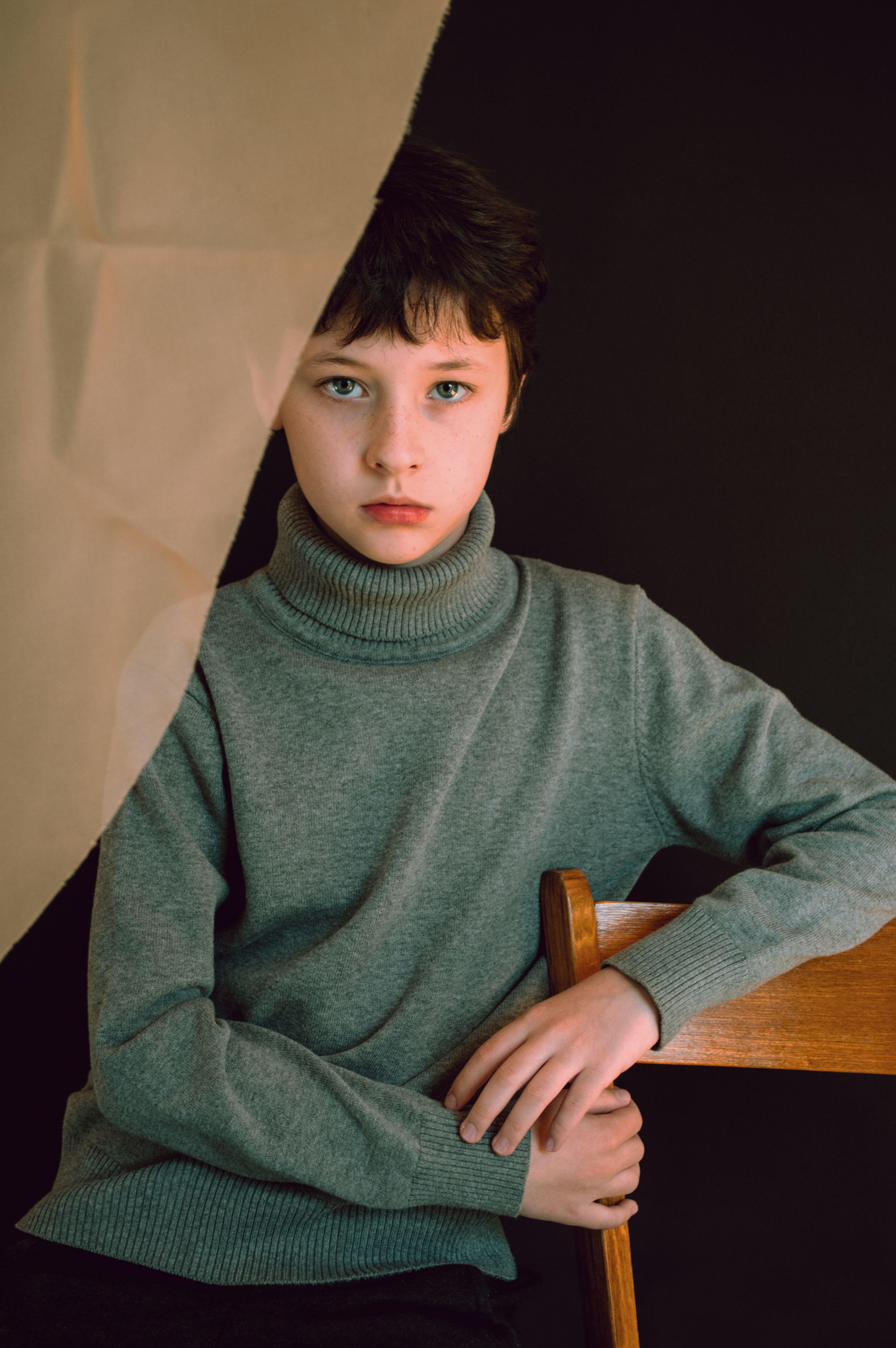 Calm male child with brown hair wearing warm gray turtleneck sweater sitting on wooden stool and looking at camera against black background