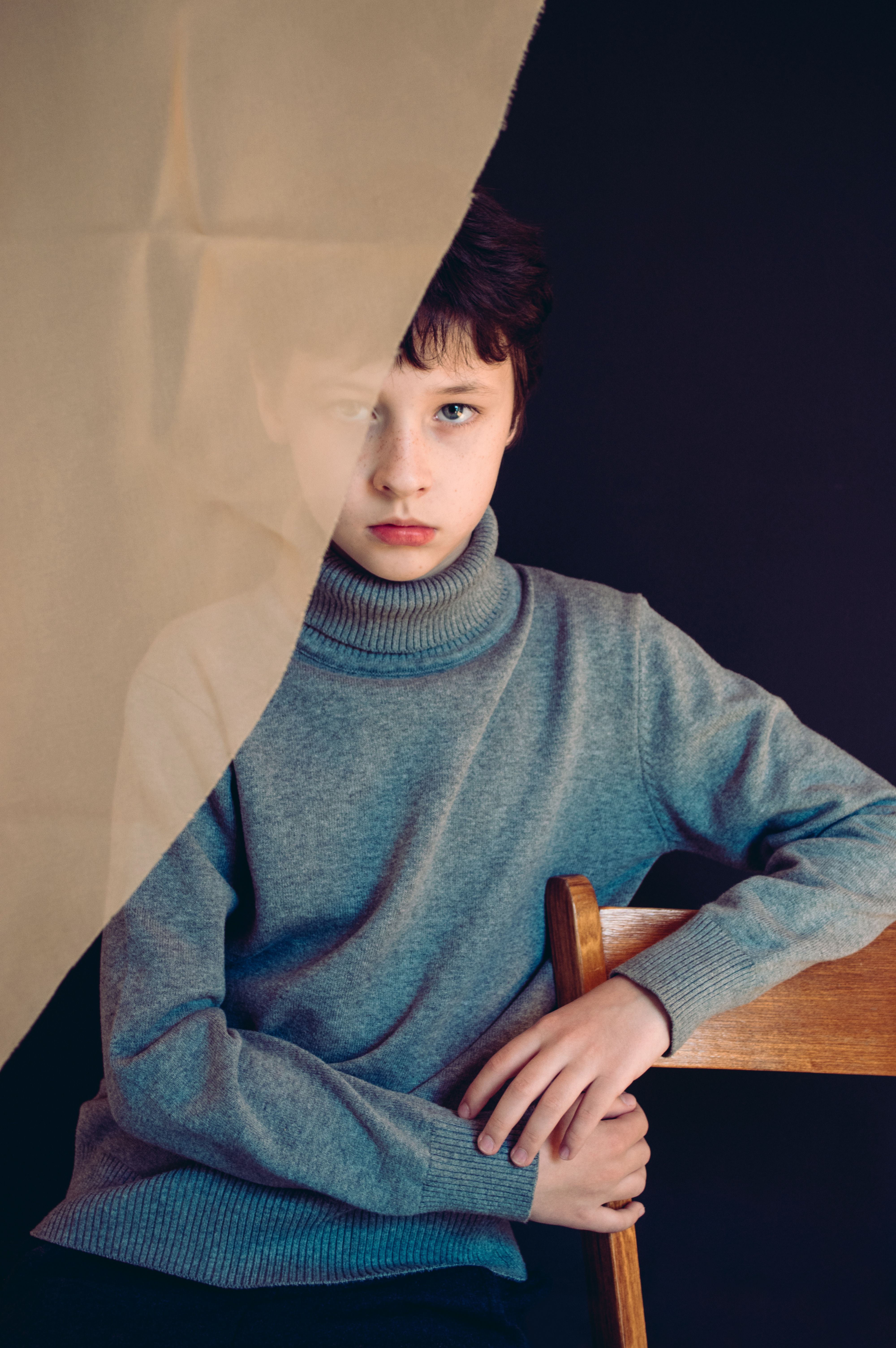 Emotionless male child with brown hair wearing warm gray turtleneck sweater covered with transparent curtain sitting on wooden stool and looking at camera against black background