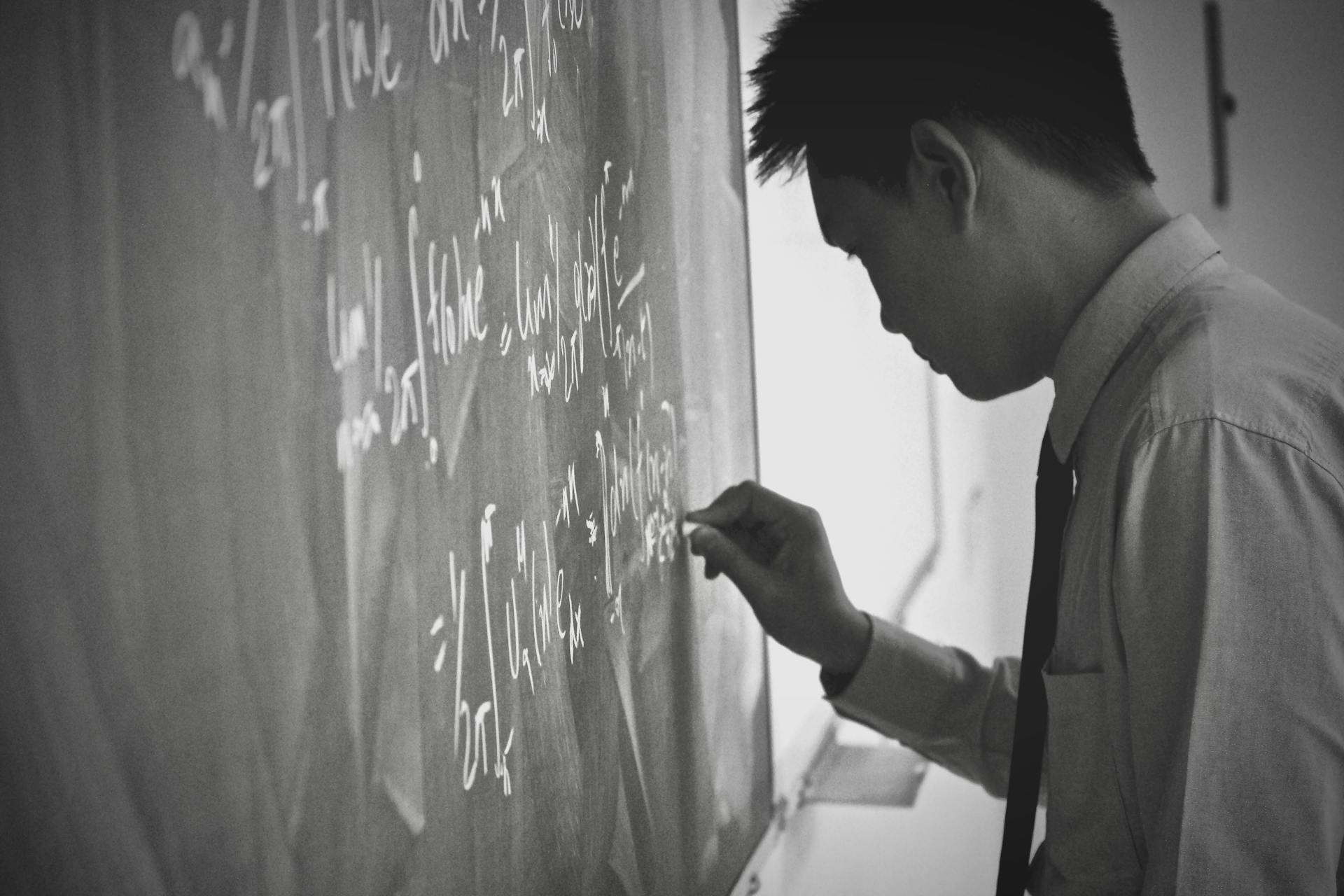 Monochromatic image of an Asian man writing math equations on a blackboard in a classroom.