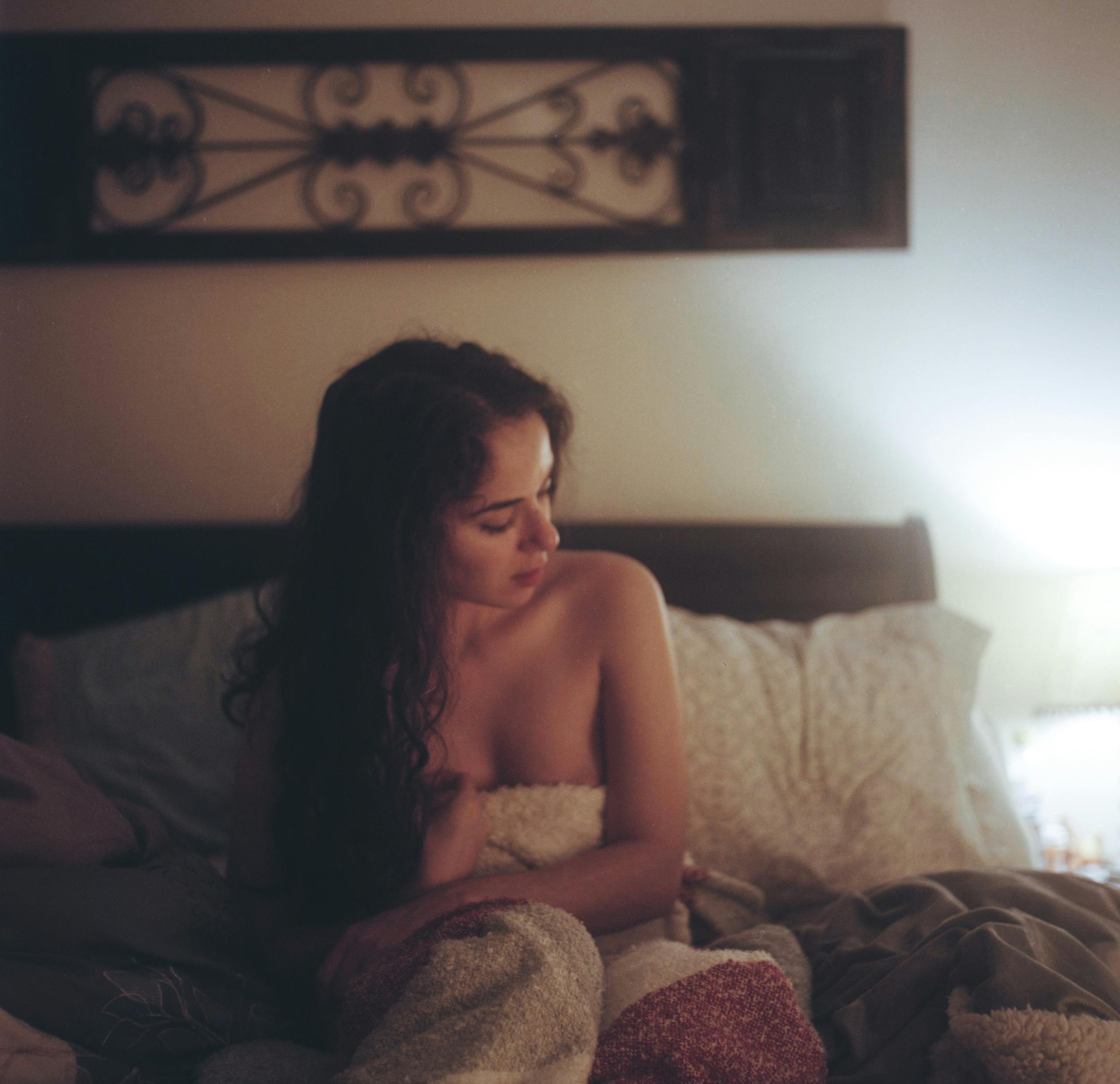 topless woman with white and red blanket on bed with pillows under brown frame inside white painted room