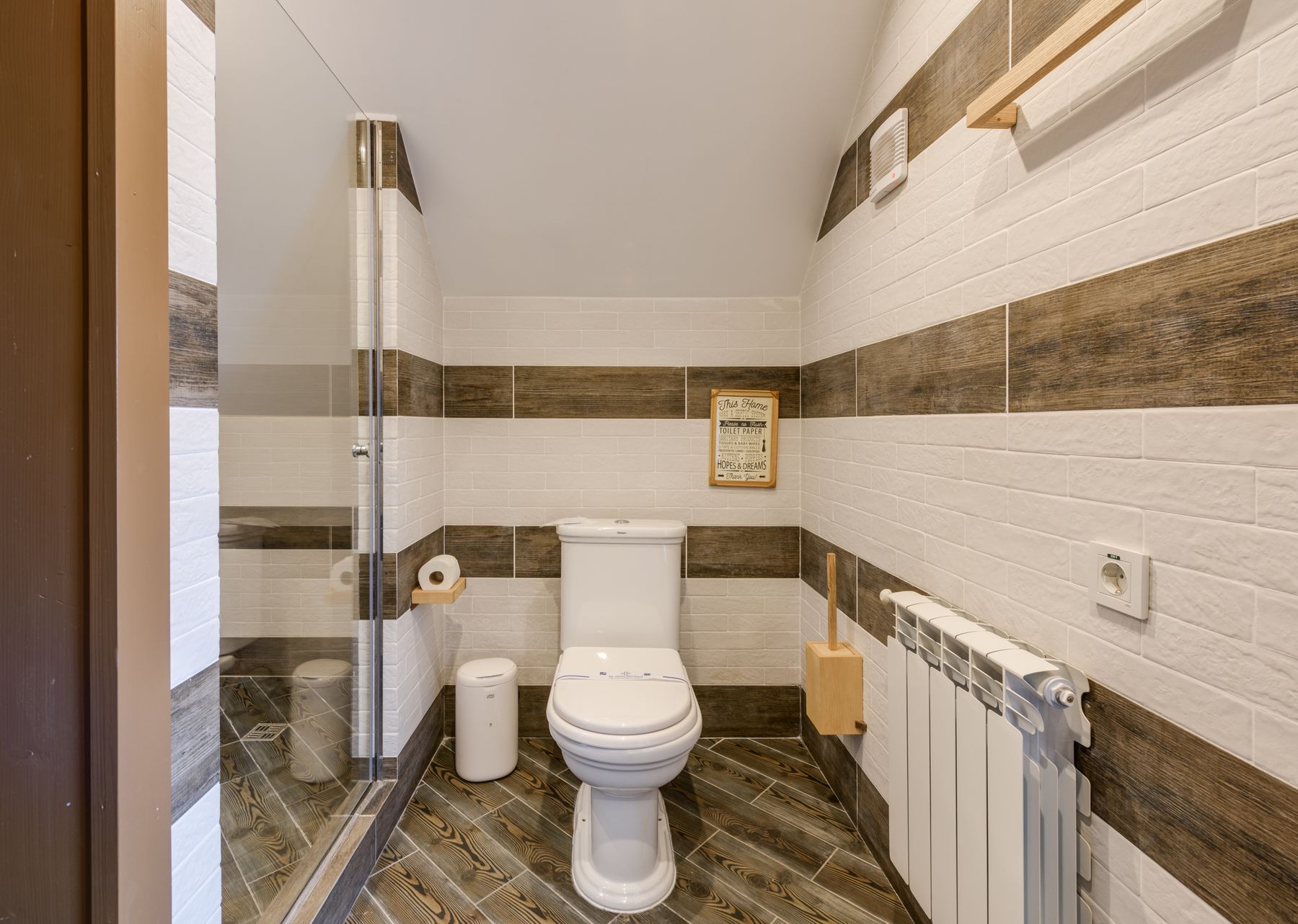 Small bright bathroom with pale tile, warm wood vanity and a clear glass shower