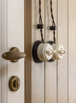 Close-up of vintage ceramic light switches with twisted wires on a wooden panel wall, featuring a brass door handle.