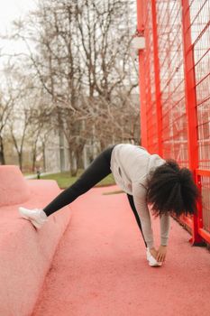 African American woman stretching outdoors by a red fence in a park setting, promoting healthy lifestyle.