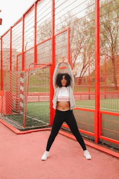 A young woman stretching in sportswear by a soccer goal at an outdoor gym with a red metal fence.