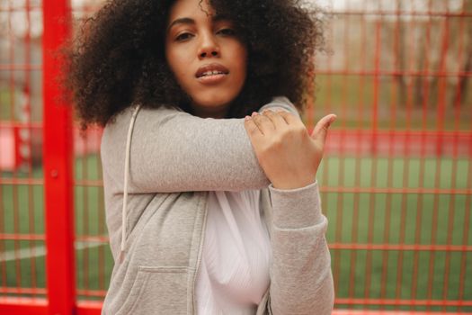 A young woman with curly hair stretches outdoors near a red fence, promoting fitness and wellness.