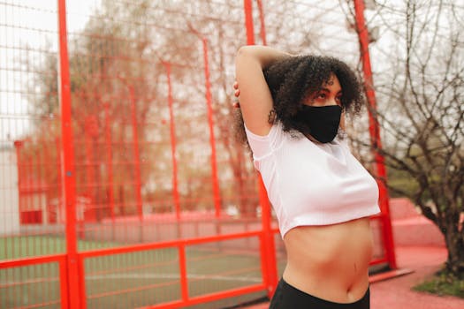 African American woman in a face mask stretching outdoors by a red sports court.