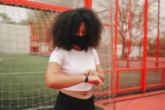 Young woman with curly hair and face mask checking time on wristwatch outdoors.