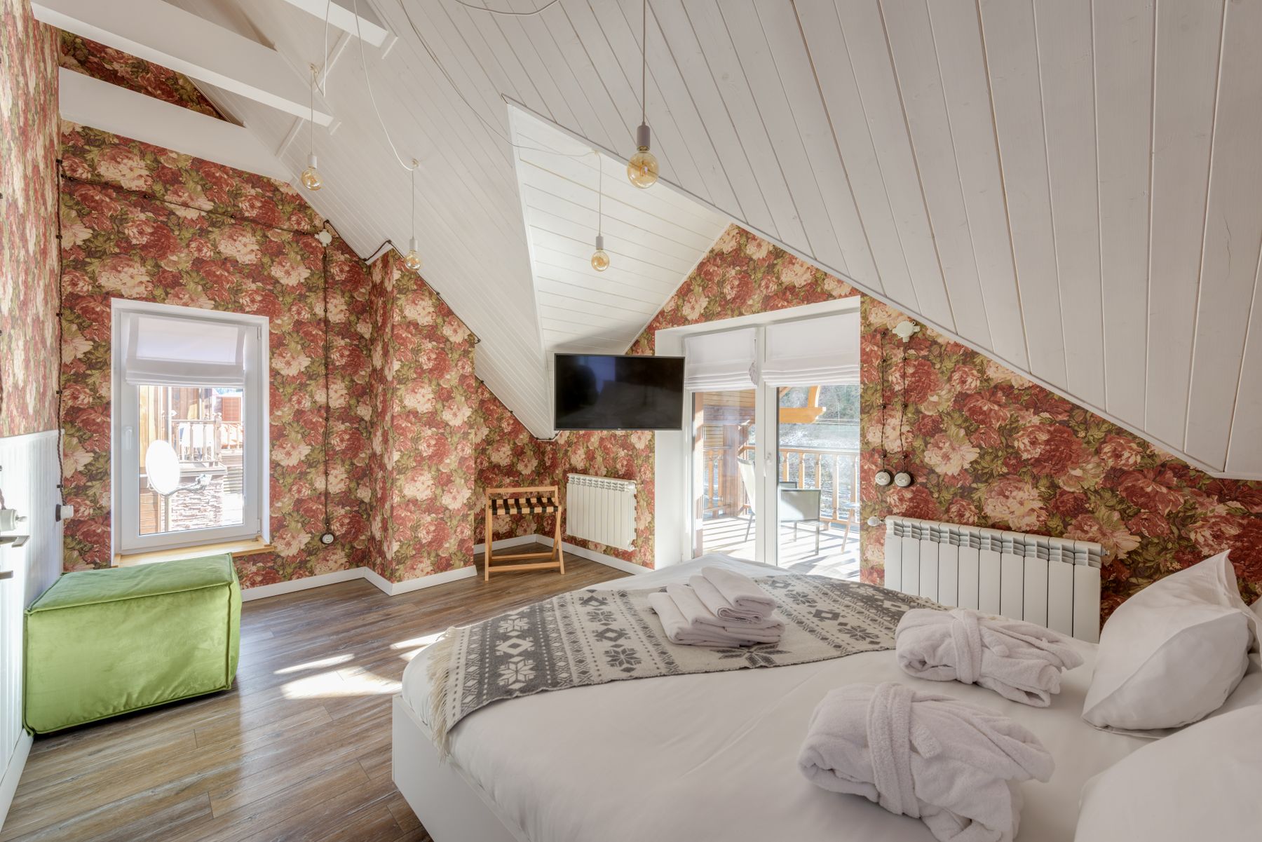 Sunlit attic country bedroom with floral wallpaper, white bed, warm timber floor, sloped ceiling and balcony doors