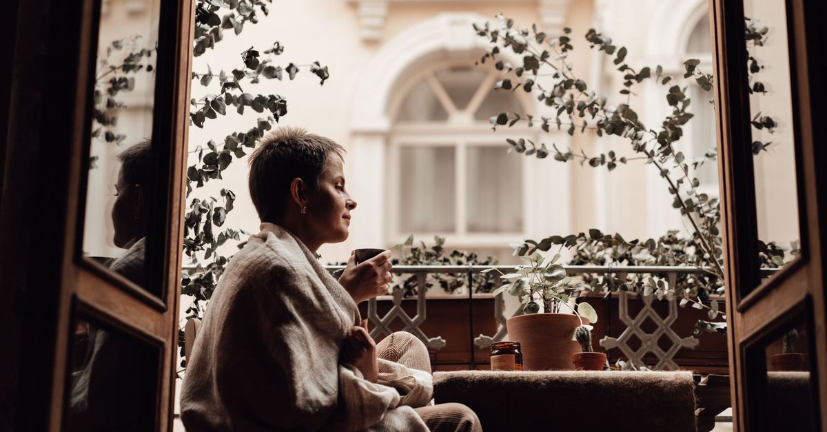 Woman wrapped in a blanket sipping tea on a charming balcony with plants.
