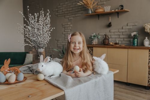 Free Delighted little girl with Easter eggs lying on table with white rabbits at home and looking at camera Stock Photo