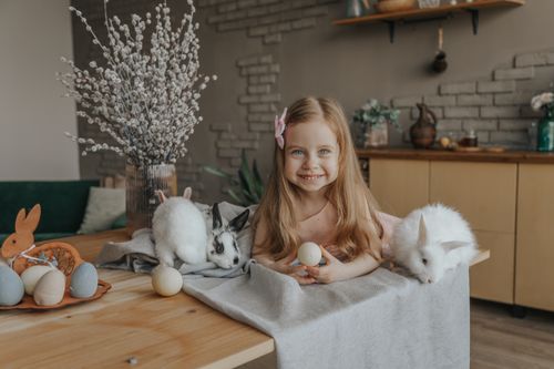 Free Positive little child lying with egg on table with cute fluffy bunnies during Easter celebration at home and looking at camera Stock Photo