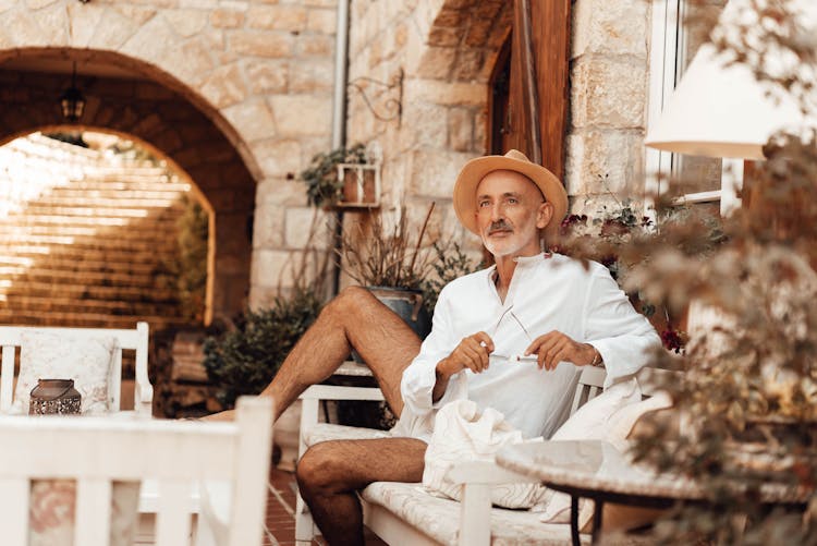 Beige Portrait Of A Man In A Summer Hat Sitting At A Table Outdoors And Holding Eyeglasses