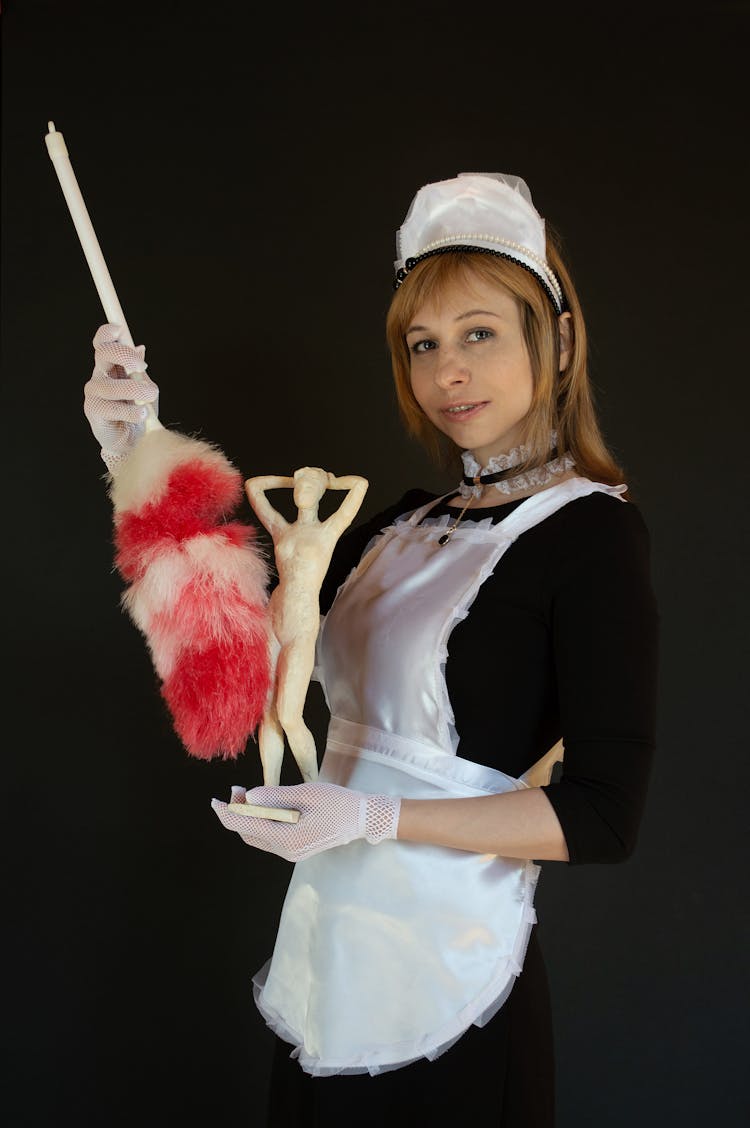 Smiling Young Woman In Uniform Of Maid Dusting Statuette