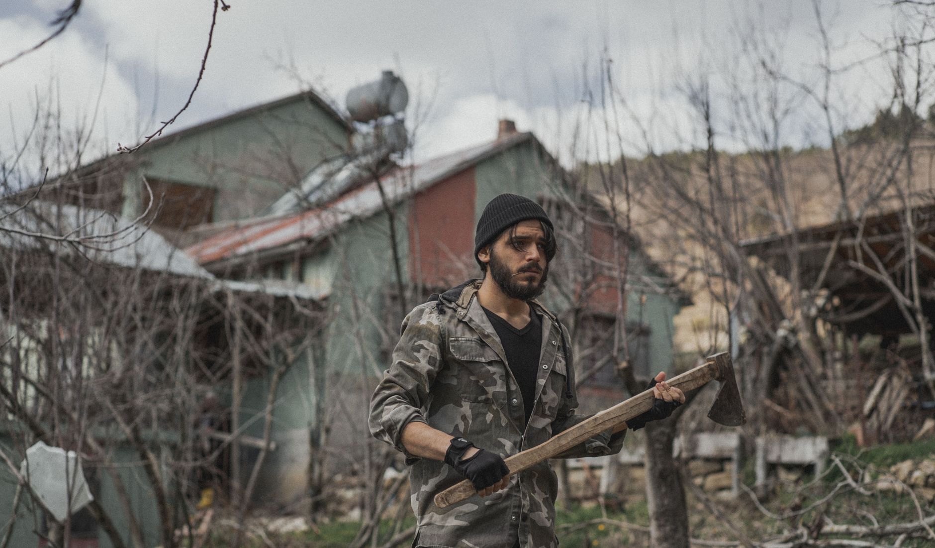 A rugged man wearing camouflage and a beanie stands outdoors with an axe, depicting survival skills.